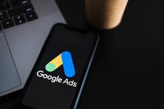 Ένα κινητό με το λογότυπο του Google Ads σε σκούρο γραφείο, δίπλα στο πληκτρολόγιο ενός φορητού υπολογιστή
